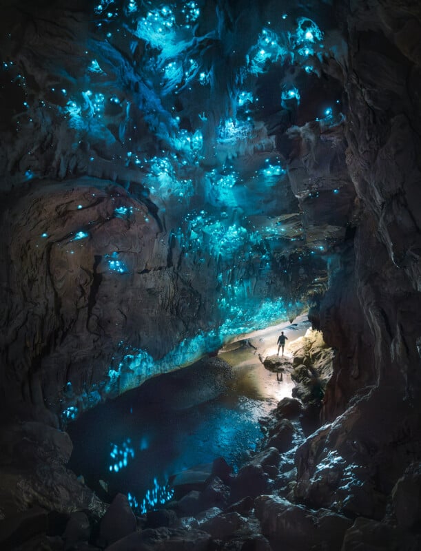 A person stands in a large cave illuminated by glowing blue bioluminescent lights on the ceiling and walls, with a small pool of water reflecting the vibrant light.