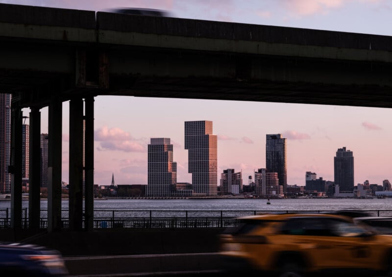 Modern city skyline with stacked, geometric high-rise buildings across a river at sunset, partially framed by an overpass and a blurred yellow taxi moving on the road in the foreground.
