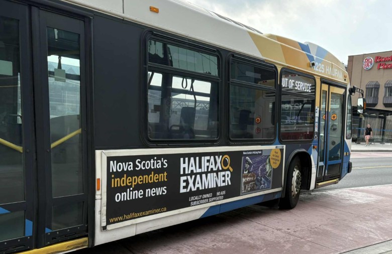 A Halifax Transit bus painted in white, blue, and yellow with a Halifax Examiner ad on the side, pulls away from a bus stop on a city street. The had says, "Nova Scotia's independent online news," with the Halifax Examiner logo with a bright yellow magnifying glass. More text on the ad says, "Locally owned, no ads, subscriber supported."