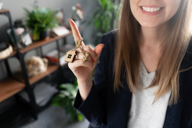 House key rotates on the finger in womans hands. Young pretty woman smiles. Modern light lobby interior. Real estate, hypothec, moving home or renting property.