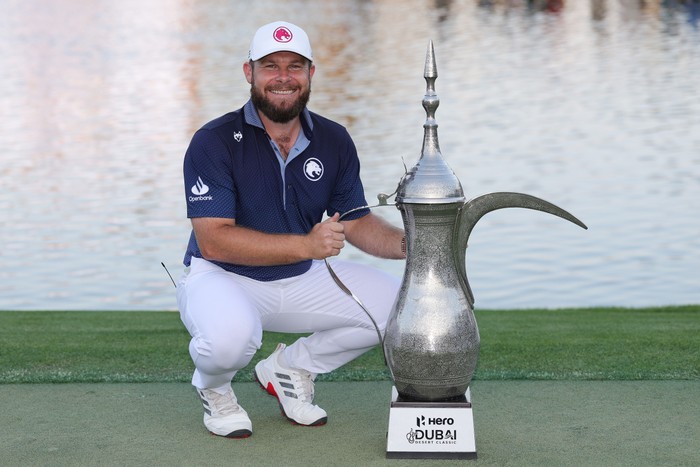 Tyrrell Hatton Tyrrell Hatton, in blue and white golf kit, smiles as he crouches with the Hero Dubai Desert Classic trophy.