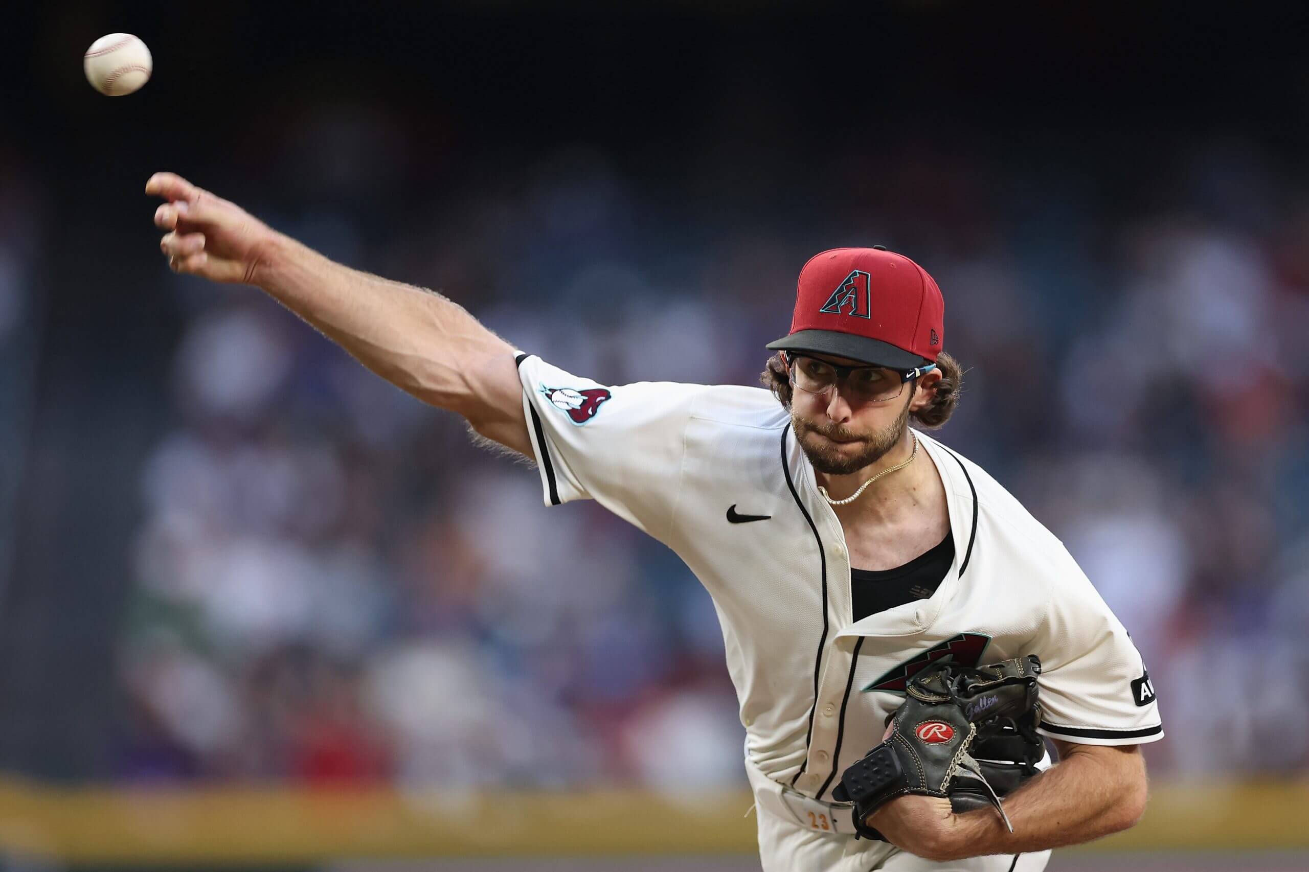 Zac Gallen, in a white jersey with a black undershirt and wearing a red Diamondbacks hat with a black bill, releases a pitch.