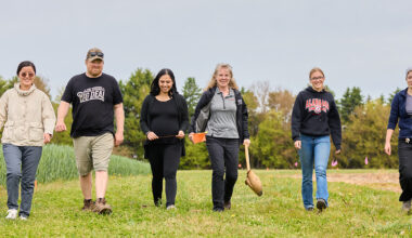 Six people walk across a grassy research field holding flags and sampling tools, with crop plots and trees in the background.