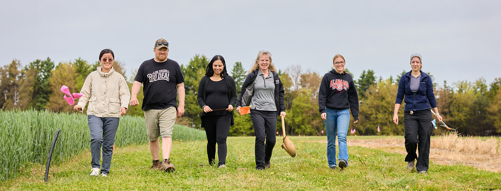 Six people walk across a grassy research field holding flags and sampling tools, with crop plots and trees in the background.