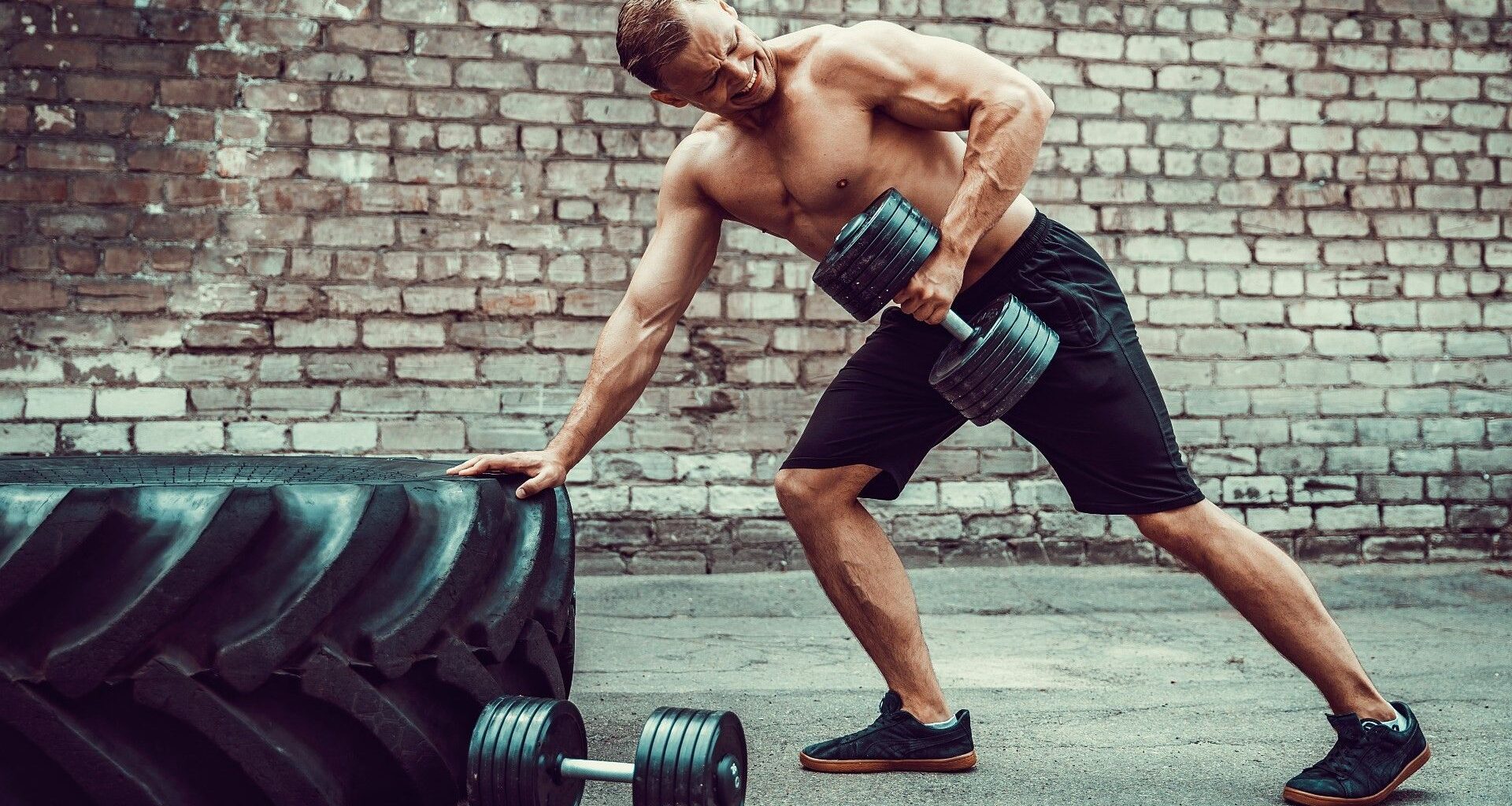 Man leaning against a tyre with one hand and performing a dumbbell bentover row with his left arm
