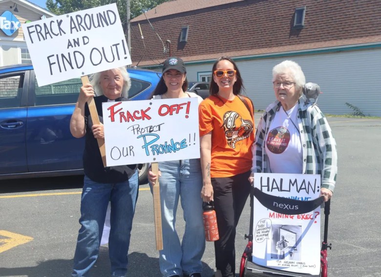 Four white woman are standing as a group in a car parking. Two women in the middle are younger and the two others on the sides are seniors. The senior woman on the far left is holding a banner stating, "Frack around and find out," on her right a young woman is holding a banner that says "Frack off! Protect our province," another young woman on her right is not holding any banner, she is wearing an orange t-shirt and orange sun glasses, on the far right is a senior woman with a walker which is carrying a banner saying Halman: mining exec.