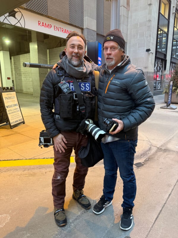 Two men dressed warmly stand on a city sidewalk, both holding professional cameras. One wears a “PRESS” vest and smiles while the other stands with a neutral expression. A building entrance and hotel sign are visible in the background.