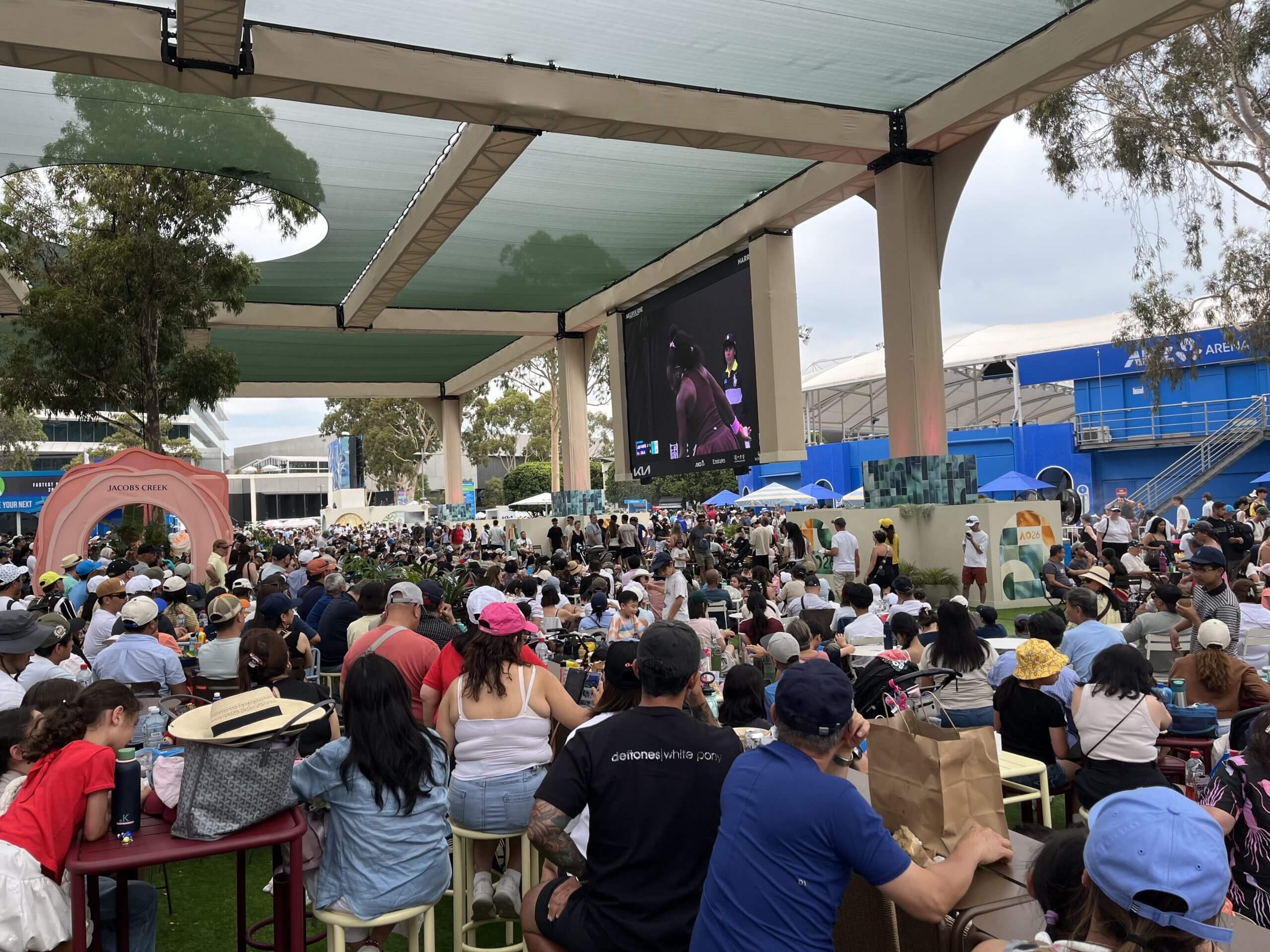 Huge crowds of fans sit on benches and the floor to watch a tennis match on a big screen at Melbourne Park.