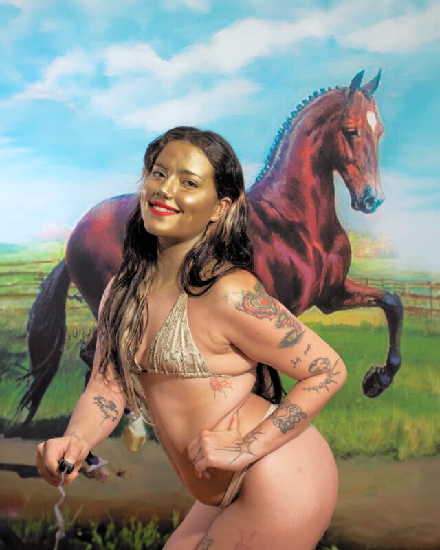 A smiling person with long hair, gold-painted face, and various tattoos poses in a bikini in front of a painting of a galloping brown horse in a grassy field under a blue sky with clouds.