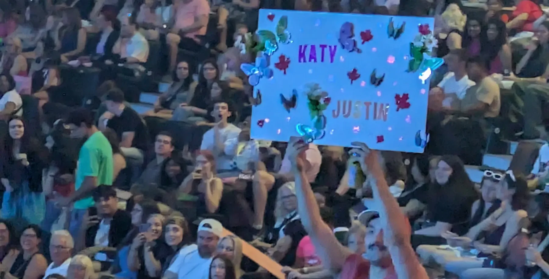 A diverse audience at a concert or event in Toronto, holding a handmade sign with illuminated decorations supporting Katy and Justin, with city-friendly keywords.