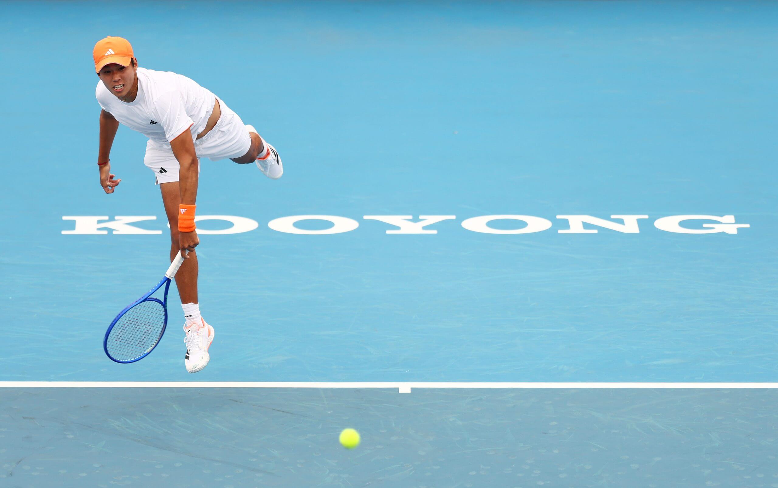 Learner Tien finishes his surface motion in mid-air on a blue tennis court, with the ball flying forward in front of him.