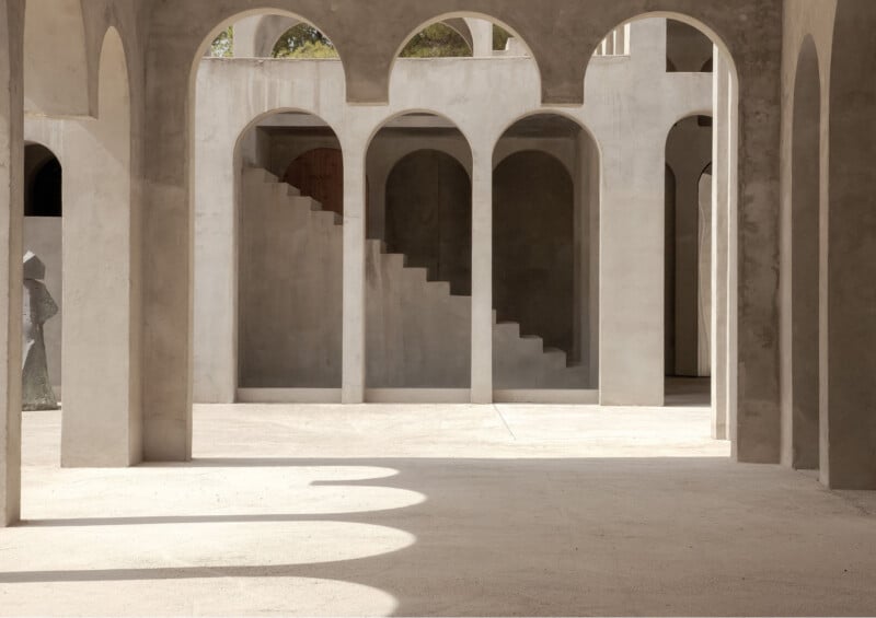 Sunlit architectural space with multiple arched doorways, columns, and a staircase leading upward in the background. The neutral-toned structure creates interesting shadows and geometric patterns on the ground.