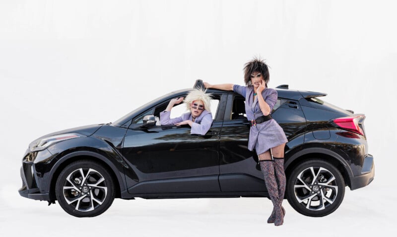 Two people dressed in bold, theatrical outfits pose with a black car on a white background; one leans out the driver’s window, and the other stands outside, resting an arm on the car roof and holding a phone.