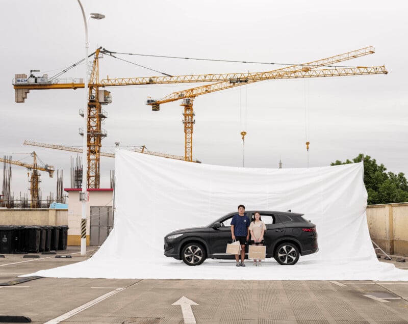 A man and woman with suitcases stand beside a black SUV, parked in front of a large white backdrop on a rooftop. Yellow construction cranes and an unfinished building are visible in the background.