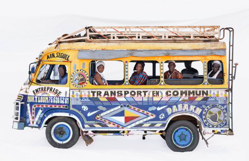 A colorful Senegalese minibus, decorated with vibrant patterns and text, has six people inside looking out the windows, posed against a plain white background.