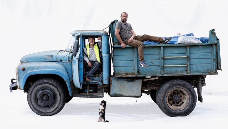 A man in a yellow vest sits in the driver’s seat of an old blue truck, while another man sits on the truck bed. A small black-and-white dog stands on its hind legs in front of the truck against a white background.