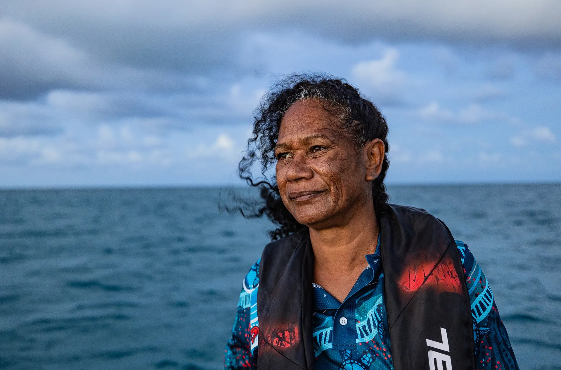 A woman with black hair and brown skin stands in front of the ocean and looks into the distance. 