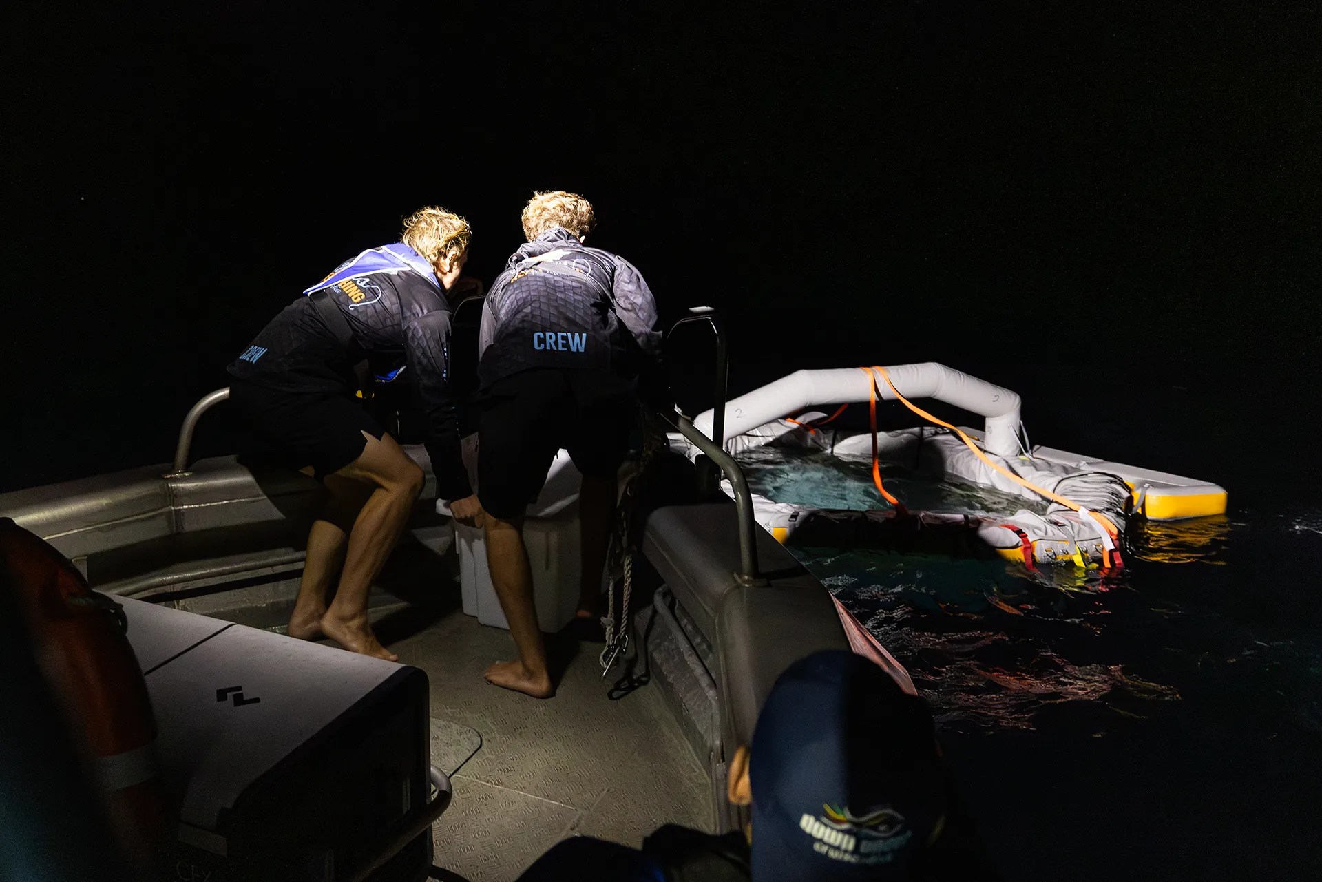 Two people hunch over the edge of a boat in the darkness.