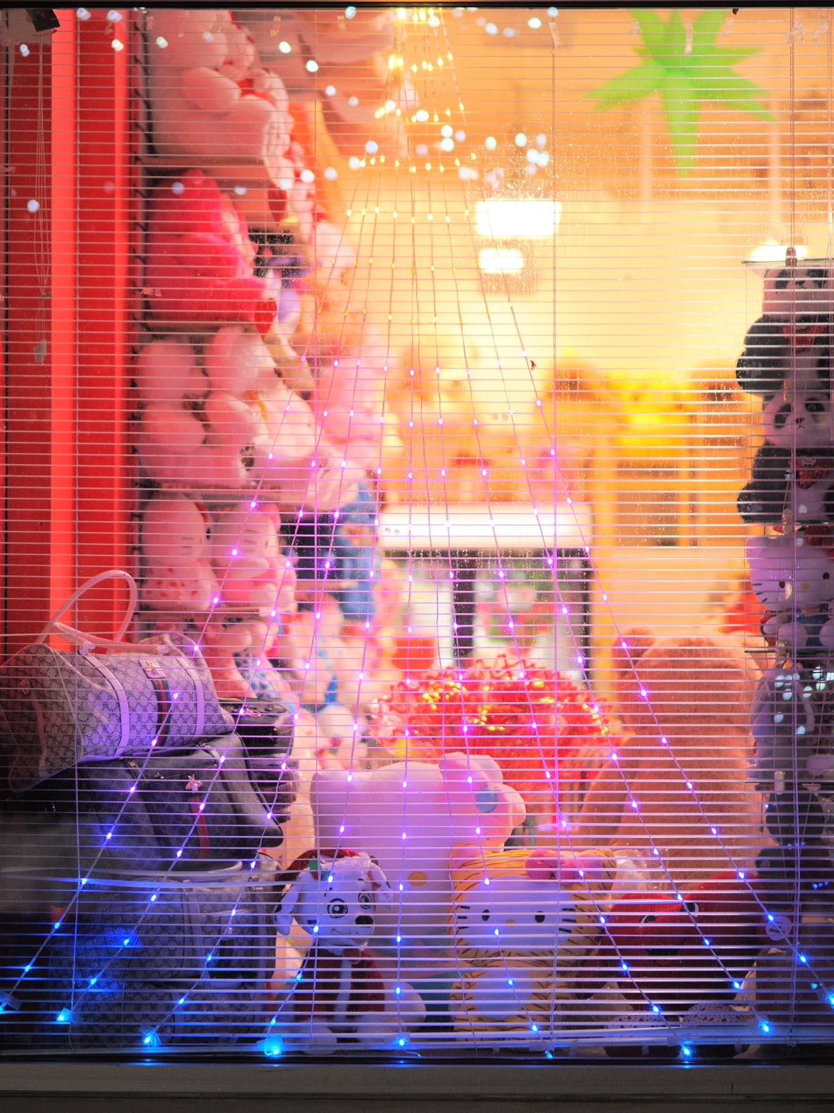 A brightly lit shop window displays plush toys, including Hello Kitty, with string lights forming a web pattern across the glass. Soft, colorful stuffed animals fill the scene, creating a warm and inviting atmosphere.