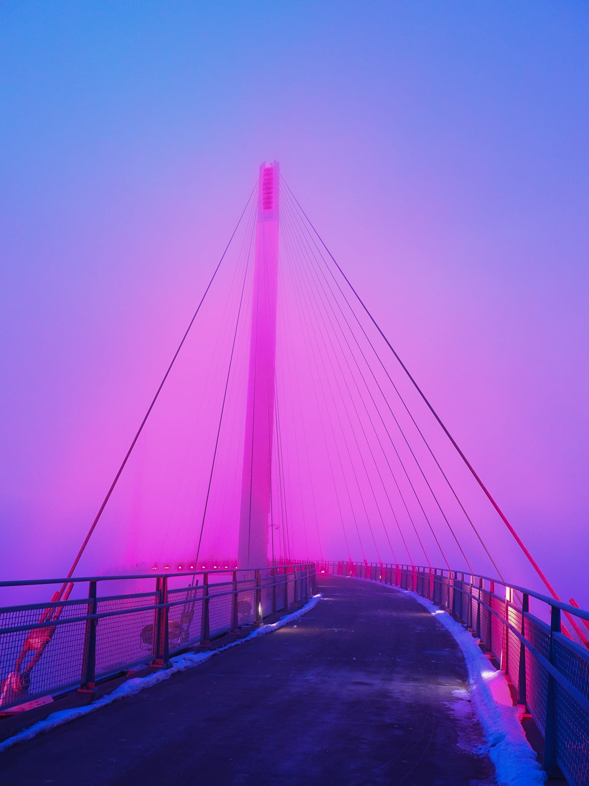 A cable-stayed bridge illuminated with vibrant pink and purple lights, shrouded in mist, with a snow-dusted pathway and railings leading toward the central tower at dusk or dawn.