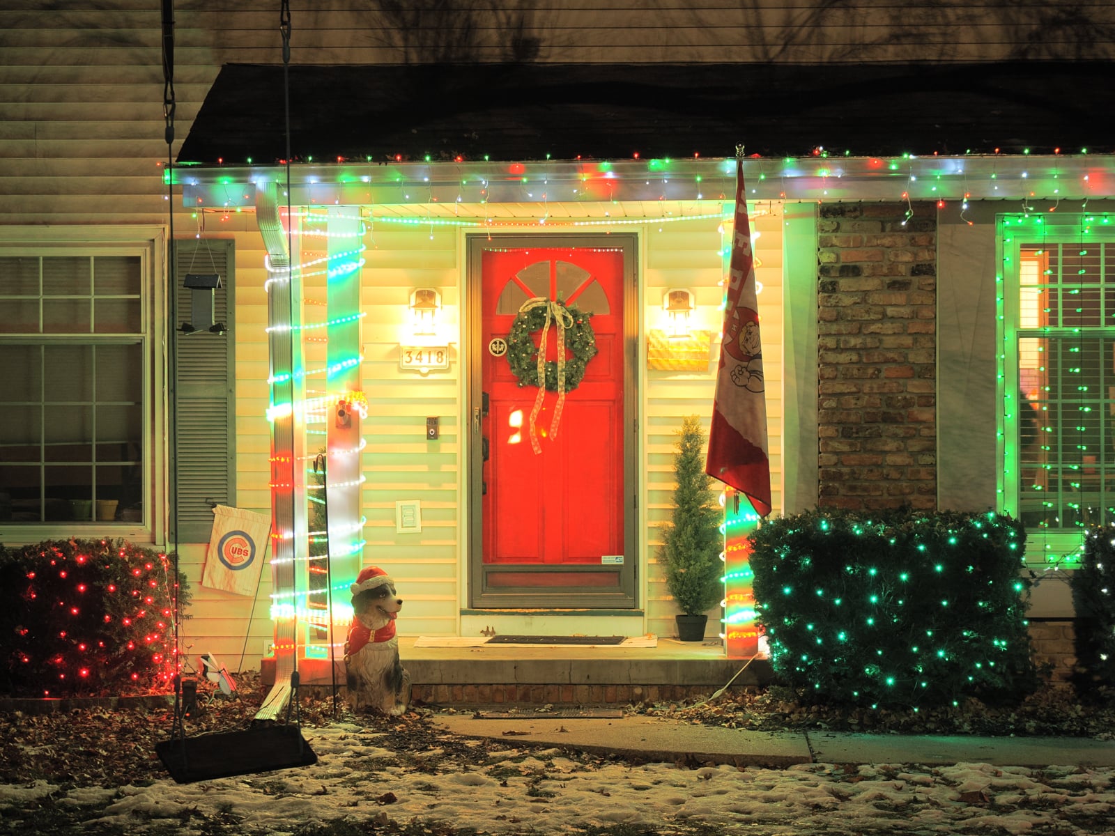 A house decorated with colorful string lights for the holidays, featuring a bright red front door with a wreath, a small dog statue in a red scarf on the porch, and a flag and shrubs nearby.