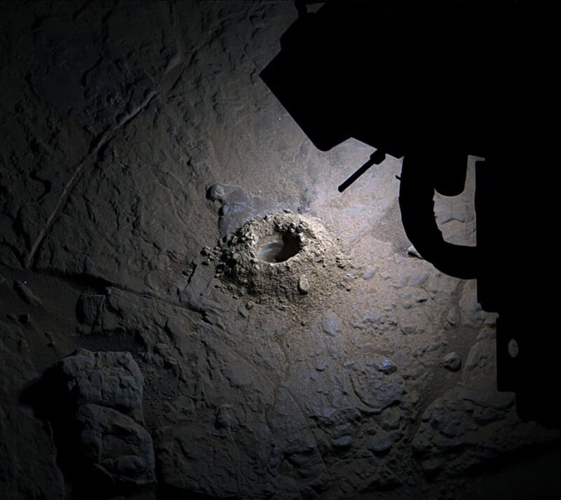 A circular hole drilled into rocky, dusty terrain is illuminated by a light source. The shadowed outline of a robotic arm or mechanical device is visible in the upper right corner of the image.