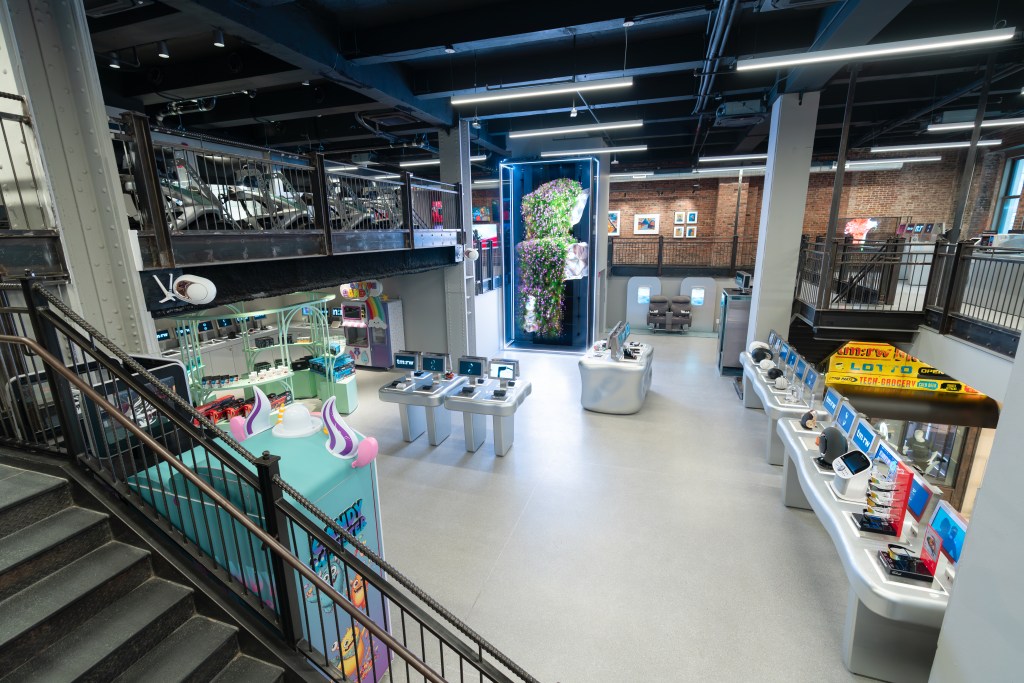 A multi-story brick-walled room with a gray floor, housing a large 3D hologram in the center and multiple silver displays of various tech devices, speakers and simulators.