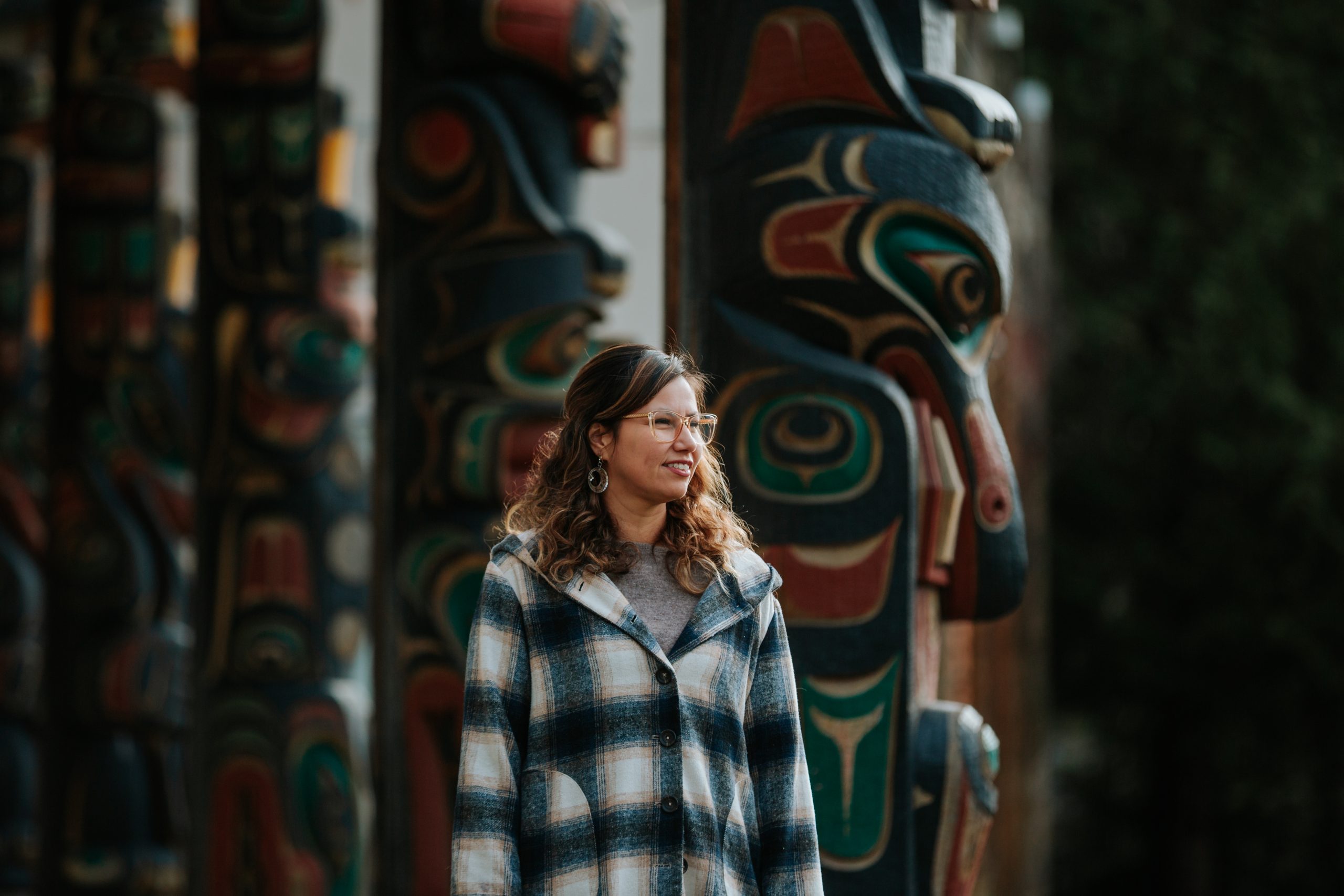 Kara Wilson stands in front of a row of totem poles, which fill the background left to right, facing the right. Kara faces to the right also, and soft sunlight illuminates the side of her face and her hair, and the edges of the totem pole figures.