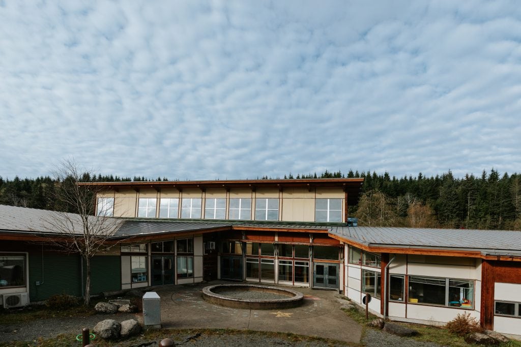 A wide view of Quatsino's school, with solar panels spanning the roof. A courtyard is centred in front of the two story building, and narrow lines of clouds line the blue sky above.