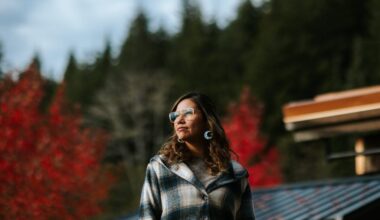 Quatsino energy champion Kara Wilson looks to the left into the soft sunlight, with wavy brown hair and lasses. Behind her, green and red trees are also aglow in the sun, and solar panels are visible on the roof of the building behind her.