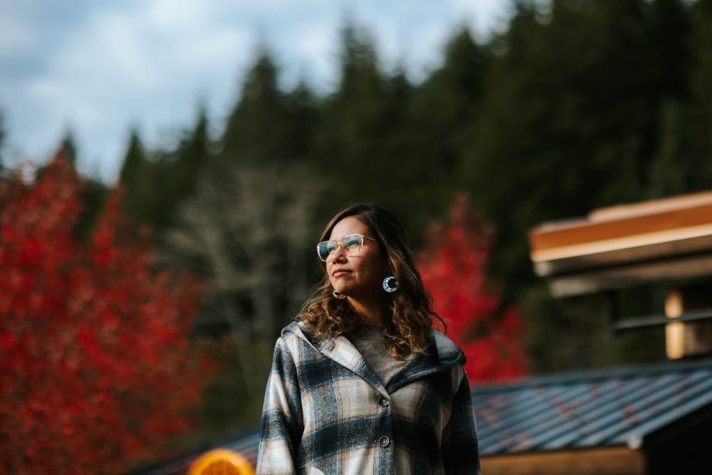 Quatsino energy champion Kara Wilson looks to the left into the soft sunlight, with wavy brown hair and lasses. Behind her, green and red trees are also aglow in the sun, and solar panels are visible on the roof of the building behind her.