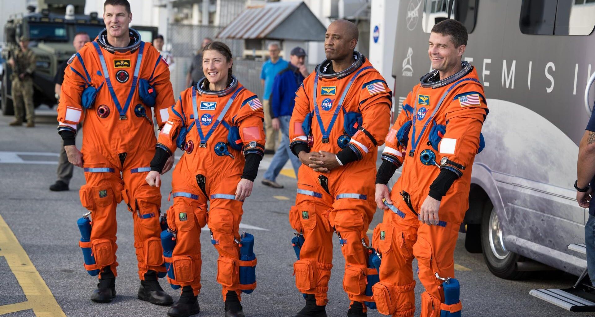 Four people in orange space suits stand on a road in front of a large van and look at a crowd off camera
