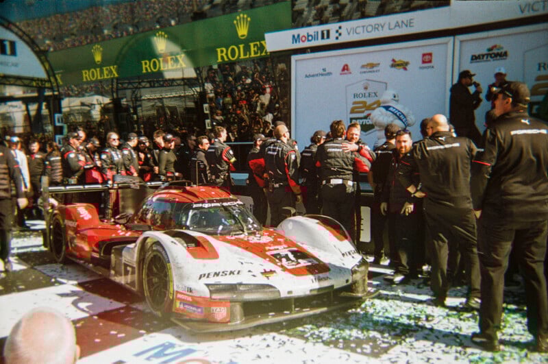A red and white Penske Porsche race car surrounded by crew and team members celebrates in victory lane at the Daytona 24 Hours event, with confetti on the ground and sponsor banners in the background.