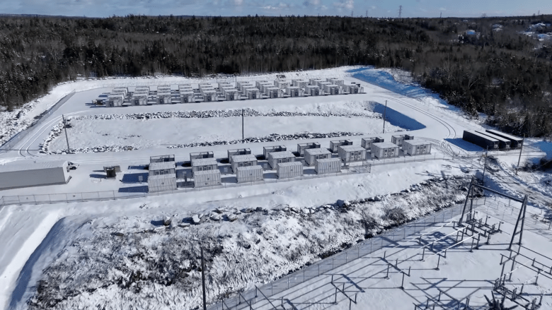 A sunny day in winter. In a large snow-covered clearing, surrounded by forest, two groupings of large white rectangular metal containers arranged in rows. One group has 29, arranged in 8 rows, the other has 87, arranged in 12 rows. Between the two groups is a shallow rectangular pit, and nearby is a gridwork of metal poles and supports.