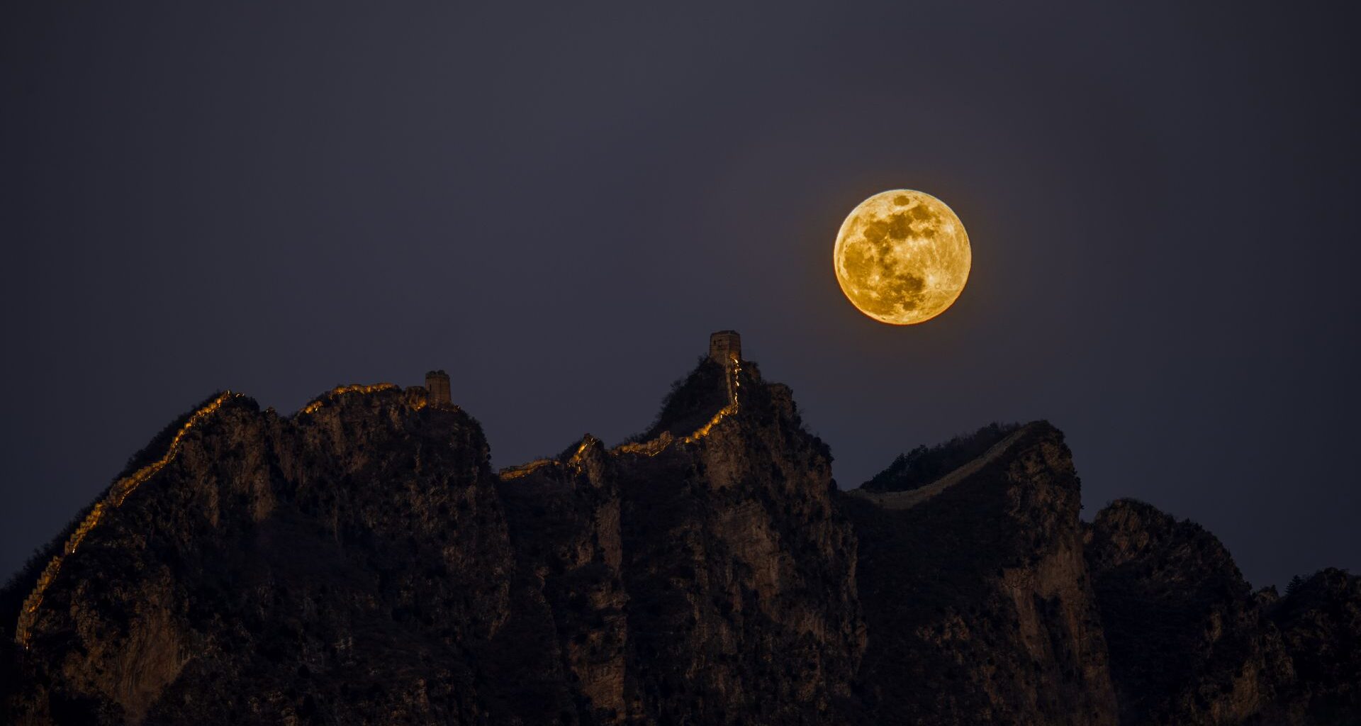 A glowing yellow full moon is seen in a gray night sky over brown jagged rocky peaks.