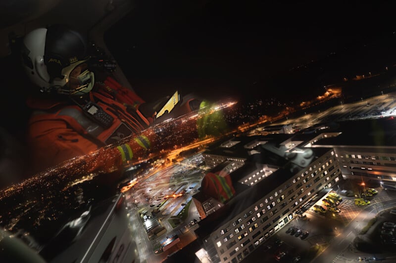 A rescue worker in a helmet and red suit sits inside a helicopter at night, using a tablet. City lights and a large hospital complex are visible below, reflected on the helicopter window.