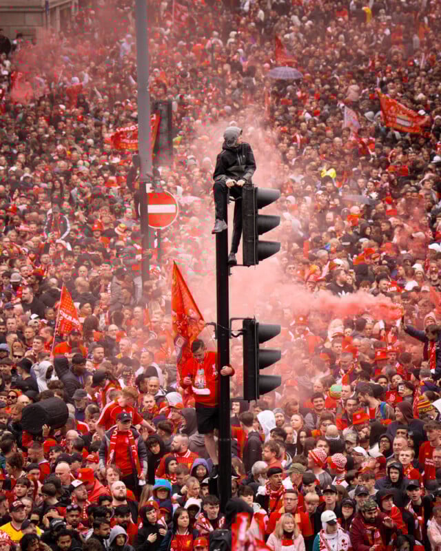A dense crowd of fans wearing red celebrate outdoors with flags and red smoke flares, while one person sits atop a traffic light above the crowd.