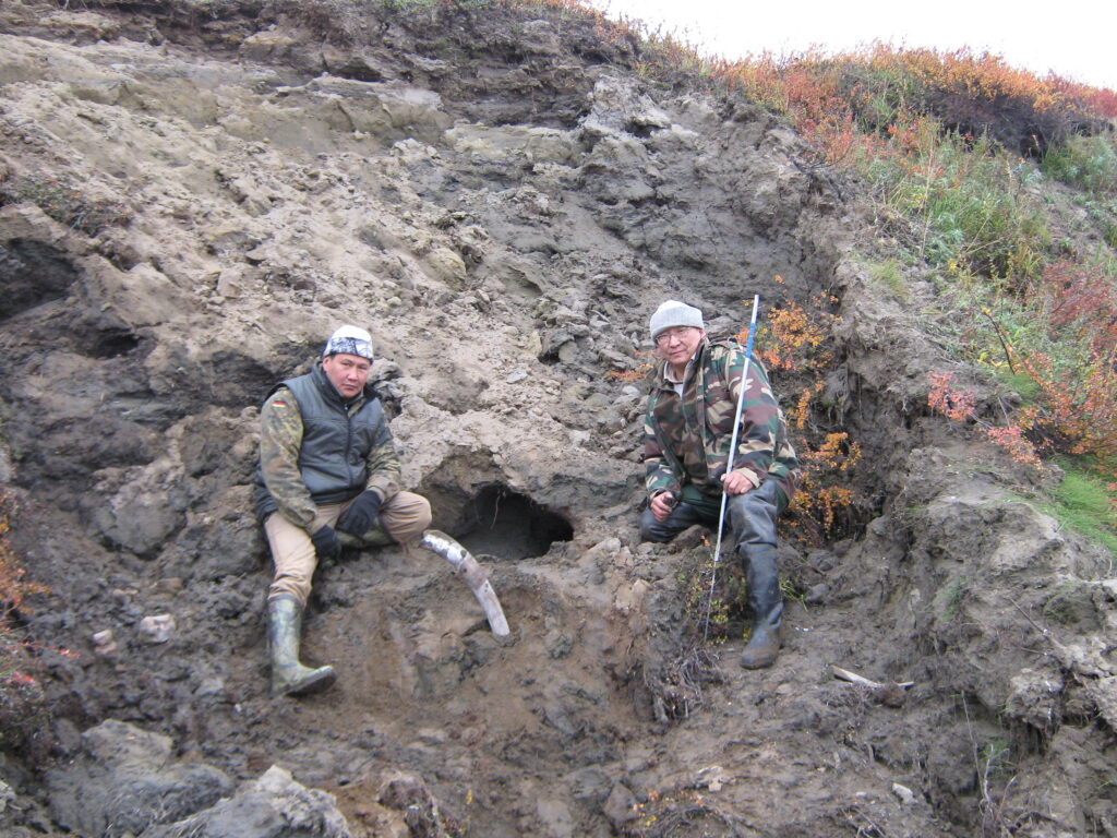 two men pose next to a hole in a muddy hillside