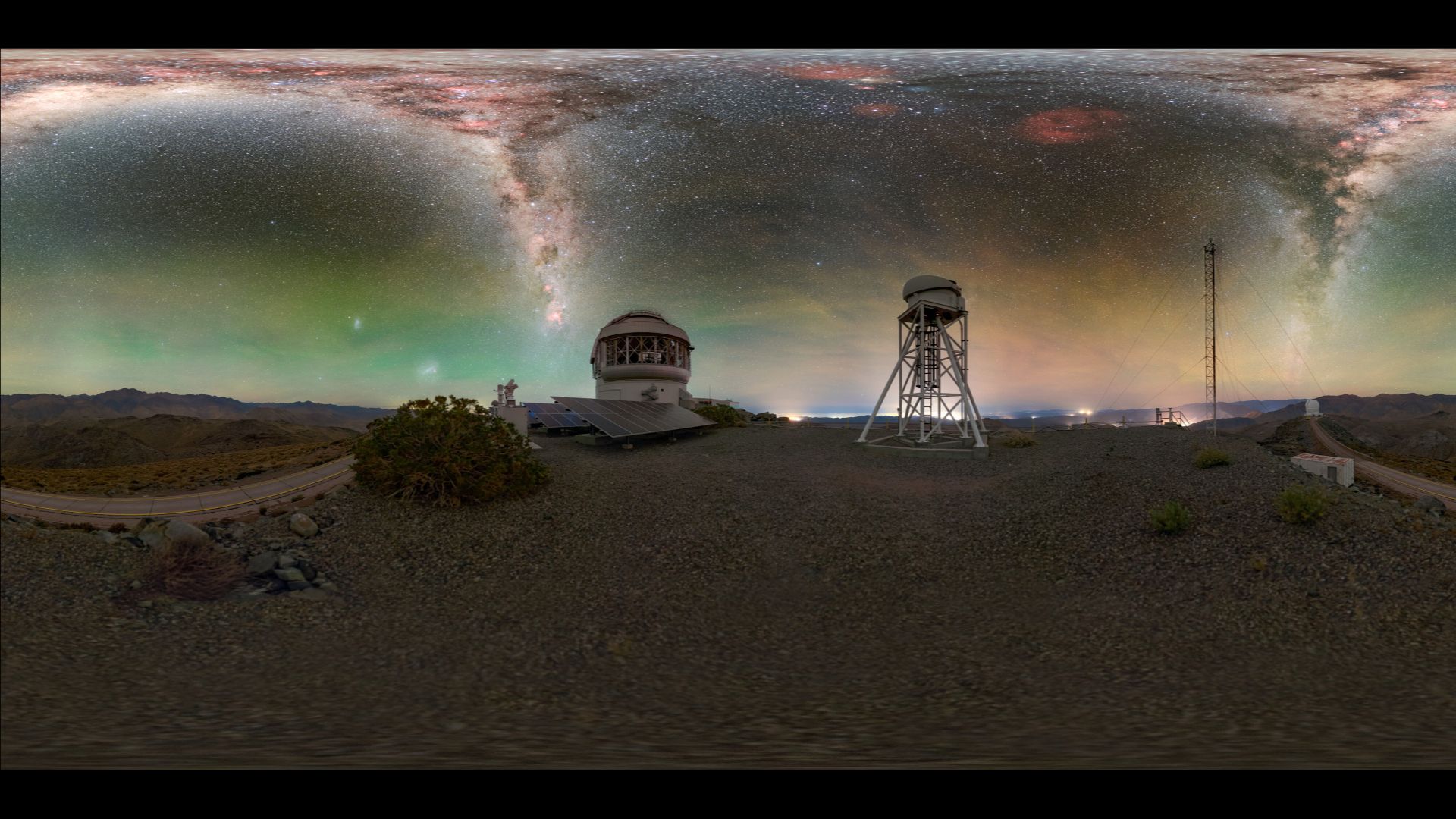 A glowing panorama shows jagged shapes of blue and purple of the Milky Way in a green and yellow night sky. Below are metal domed buildings with a barren landscape around them