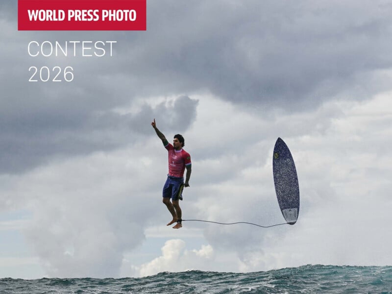 A surfer in midair points upward with one hand, while his surfboard, tethered by a leash, floats beside him above the ocean waves under a cloudy sky. Text reads "WORLD PRESS PHOTO CONTEST 2026.