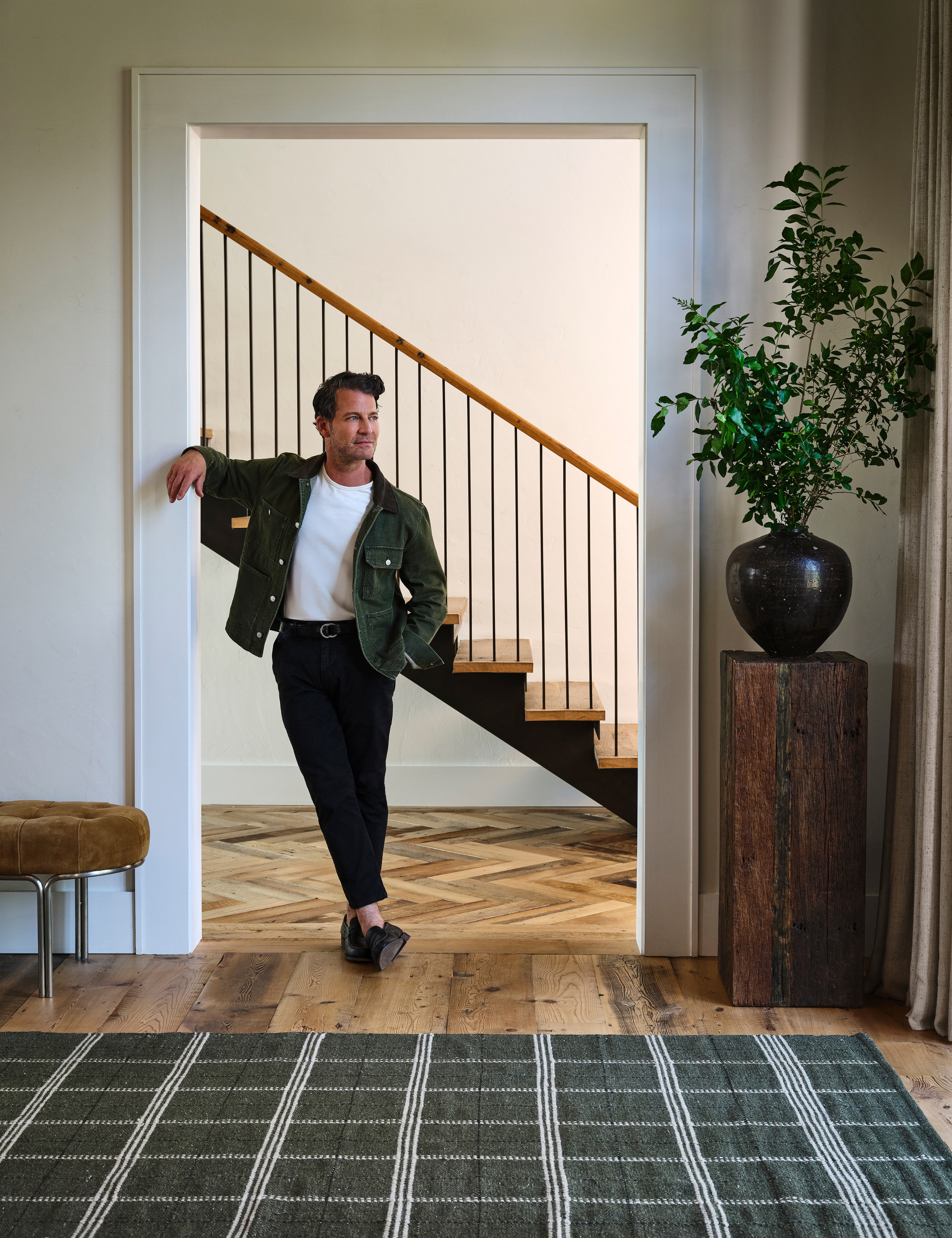 Nate Berkus posing in a classic entryway that features an evergreen plaid area rug and natural wood plinth