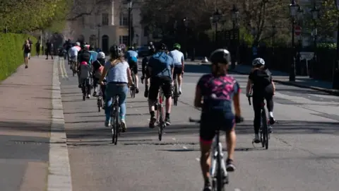 PA Members of the public cycling round Regent's Park, London. Cyclists in helmets travel along a stretch of road on a warm looking day. 