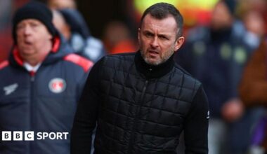 Head coach Liam Rosenior gesticulates to his players as he takes a Chelsea training session, with his arm outstretched. Fellow coach Calum McFarlane stands behind with a bunch of orange footballs, standing on one, both wearing Chelsea branded tracksuits of black beanie hats, great coats and black tracksuit trousers