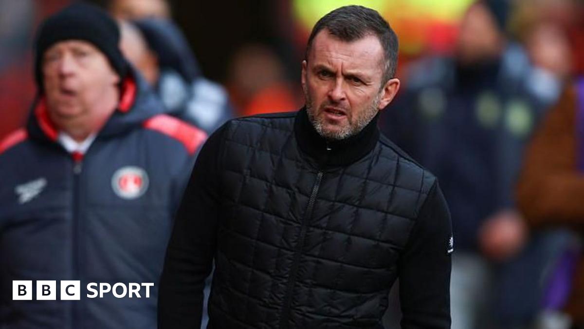 Head coach Liam Rosenior gesticulates to his players as he takes a Chelsea training session, with his arm outstretched. Fellow coach Calum McFarlane stands behind with a bunch of orange footballs, standing on one, both wearing Chelsea branded tracksuits of black beanie hats, great coats and black tracksuit trousers