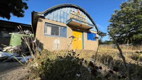 Stuart Woodward/BBC The Lifstan Boys Club building, surrounded by overgrown grass and weeds, with an overflowing green bin to the side. The building has a pebble-dashed front with two blue-framed windows and a yellow front door. The upper half of the building is a domed curved roof with blue metal fronting and the words "Lifstan Boys Club" in yellow writing.