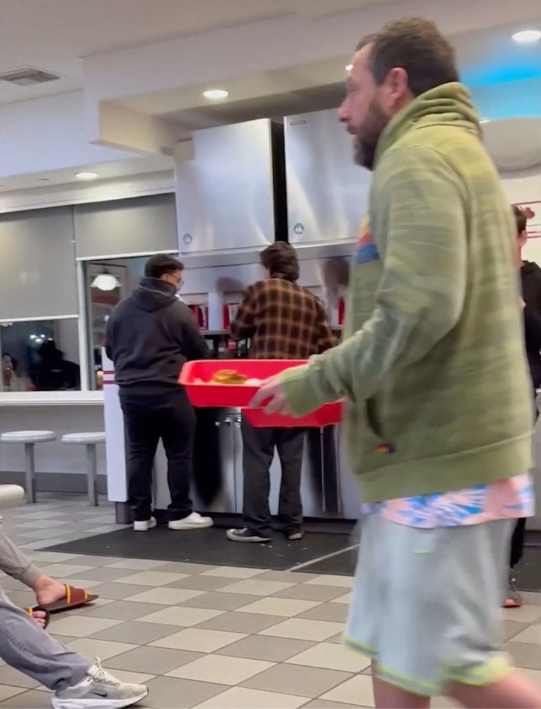 Adam Sandler walking through a fast-food restaurant with a red tray of food.