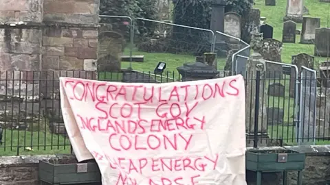 A white banner with red writing, saying Congratulations Scot Gov England's Energy Colony, hangs from railings in front of a graveyard
