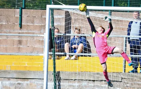 Queen of the South A goalkeeper in a pink strip makes a great save to tip the ball over the crossbar while a few people watch from behind the goals