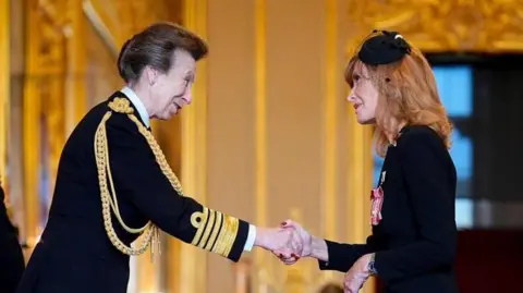 PA Media The Princess Royal shakes hands with Jackie Bird during the ceremony inside Windsor Castle. The princess is wearing a formal black navy uniform with gold braids. Jackie Bird is wearing a black suit and a black fascinator. The walls are golden