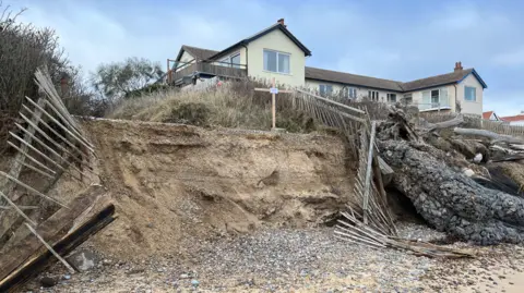 Richard Daniel/BBC An image taken from Thorpeness beach showing coastal erosion in front of a large home. Wooden fences are town down and hanging off the cliff edge.